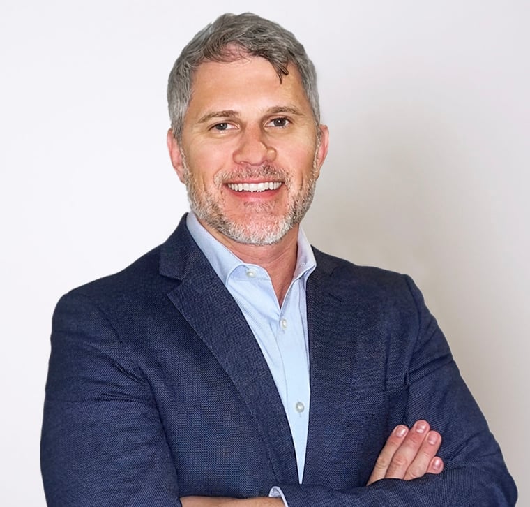 Smiling man with gray hair and beard wearing a blue suit jacket and light blue shirt, standing against a plain white background with arms crossed.
