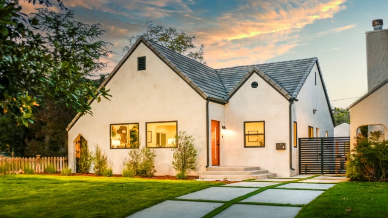 A modern stucco house with a gray roof, large windows, and a wooden front door. It has a green lawn, a small front garden, and a concrete path leading to the entrance at sunset.
