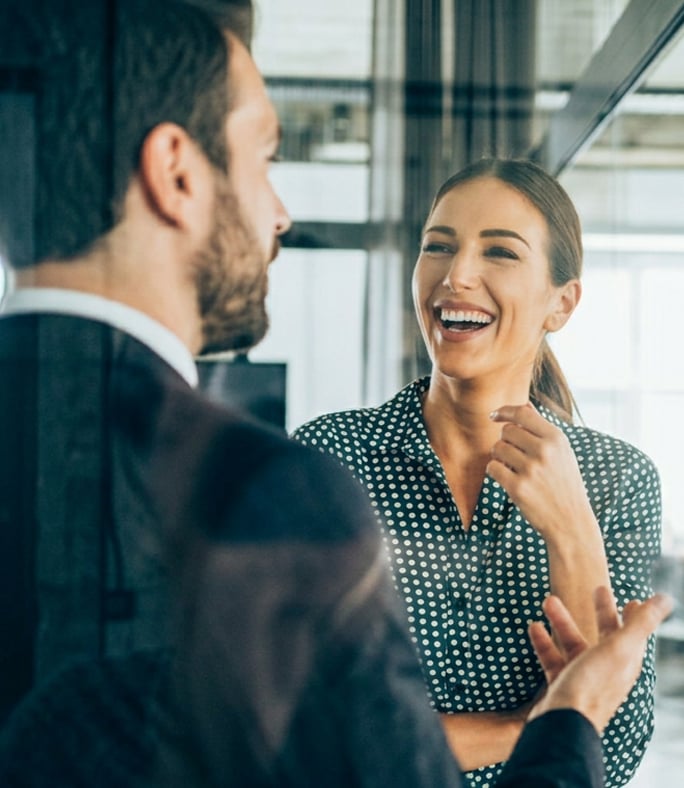 A woman in a polka dot shirt smiles while conversing with a man in a suit inside a modern office with glass walls.