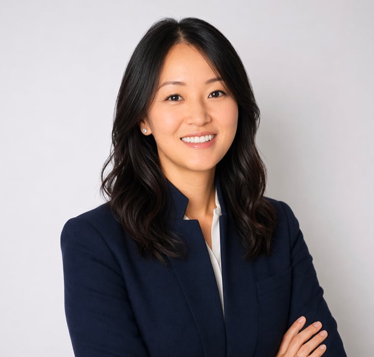 Woman with long dark hair wearing a navy blazer and white shirt, standing in front of a plain light background with arms crossed, smiling at the camera.