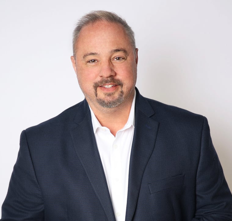 A man with short gray hair and a trimmed beard wears a dark suit jacket and white shirt, standing against a plain white background, smiling at the camera.