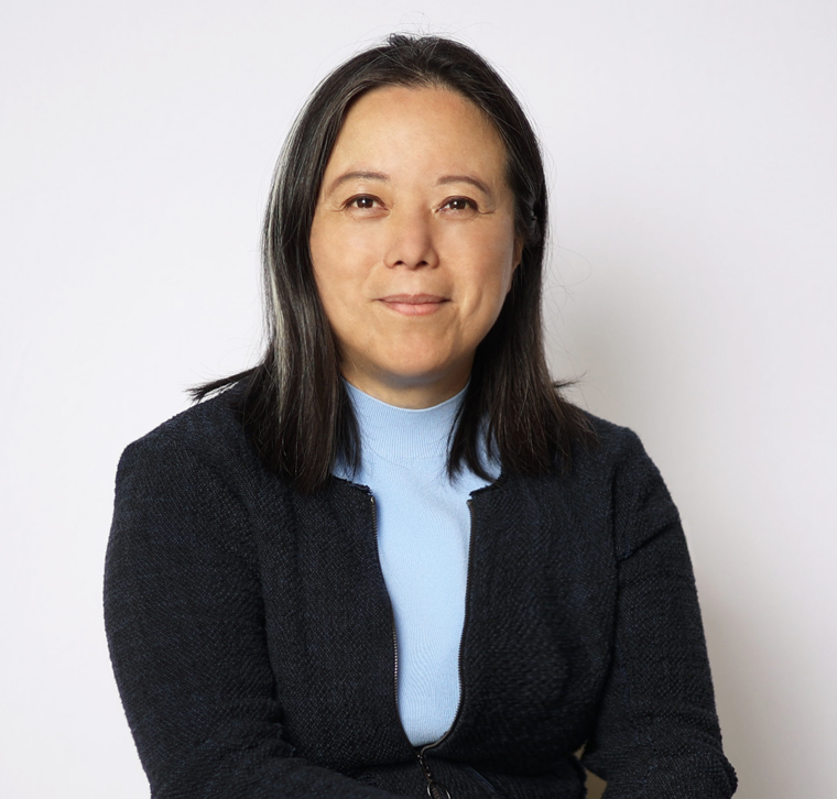 A woman with straight dark hair, wearing a blue top and dark blazer, sits against a plain light background, looking at the camera with a neutral expression.