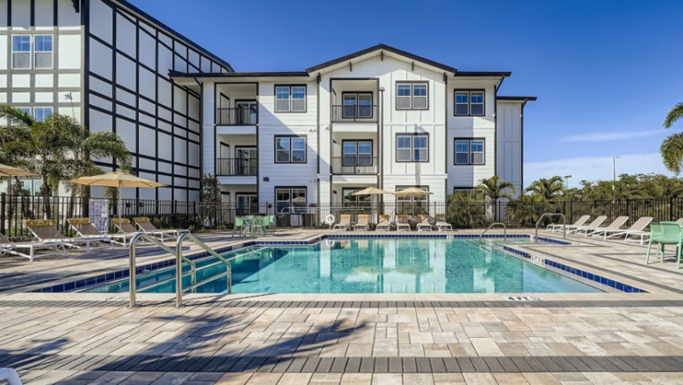 Rectangular outdoor swimming pool with lounge chairs, umbrellas, and a fenced patio area in front of a three-story apartment building under a clear blue sky.
