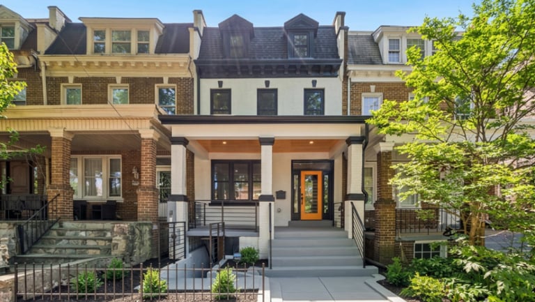 Three-story rowhouse with a black roof, white facade, orange front door, covered porch, and landscaped front yard, flanked by similar neighboring houses.