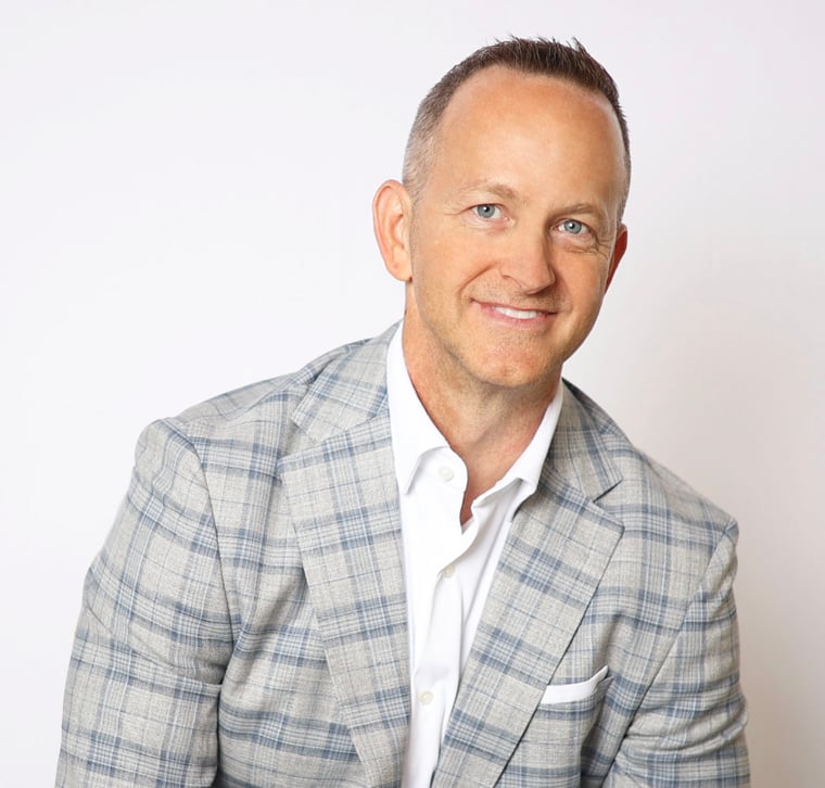 Man wearing a light gray plaid suit jacket and white shirt, smiling, seated against a plain white background.