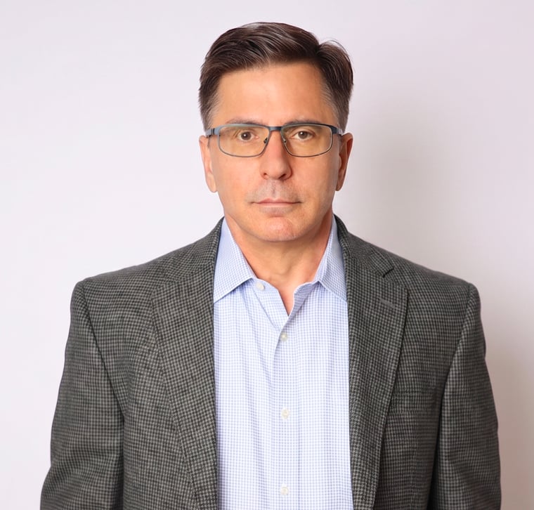 Man in glasses wearing a gray checked blazer and light blue shirt, standing against a plain white background, facing forward with a neutral expression.