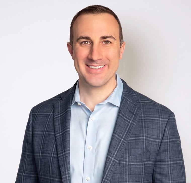 A man in a blue plaid suit jacket and light blue shirt smiles at the camera against a plain white background.