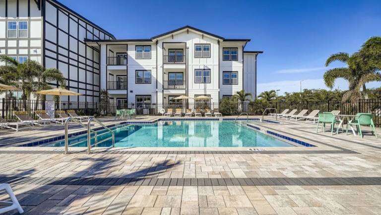 Outdoor swimming pool with lounge chairs, umbrellas, and a modern three-story apartment building in the background under a clear blue sky.