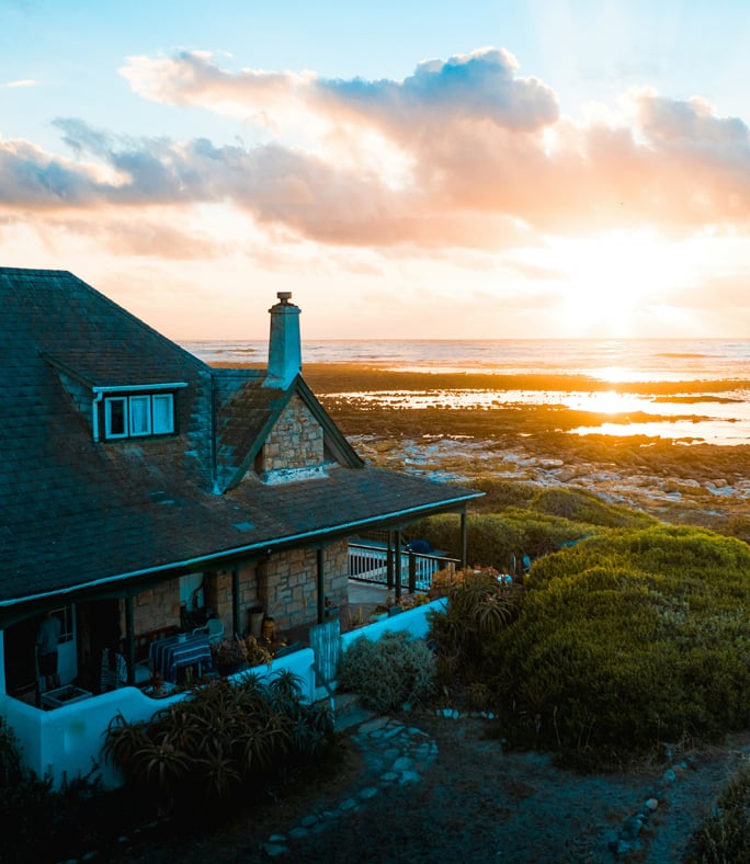 Stone house with a porch overlooking a rocky coastline at sunset, with sunlight streaming through clouds over the ocean.