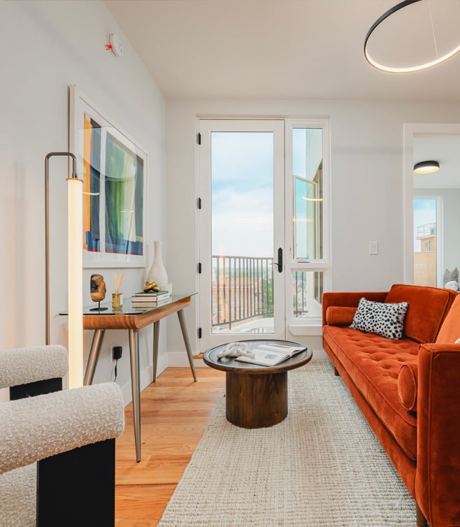 Modern living room with an orange sofa, round coffee table, and console table with decor, next to glass doors leading to a balcony. Light wood flooring and abstract artwork on the wall.