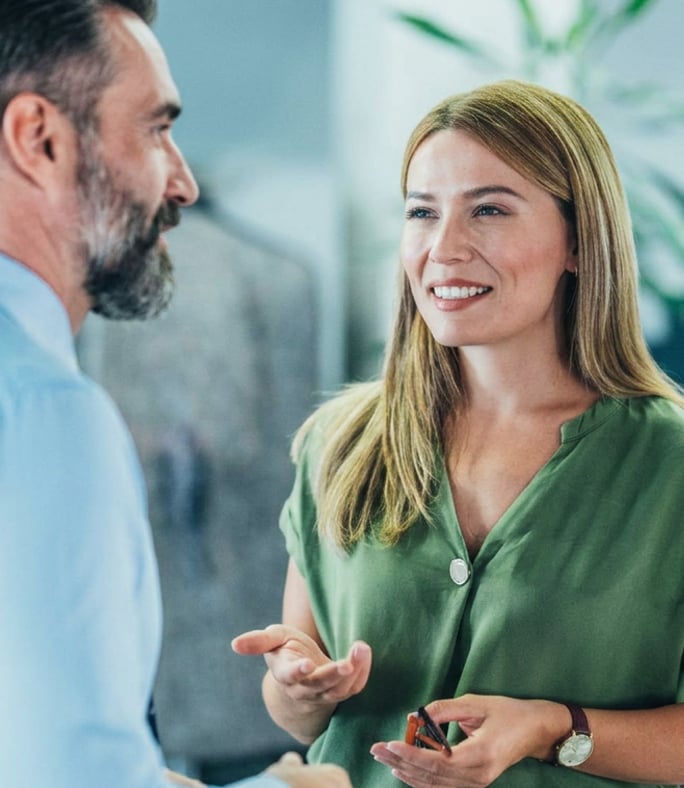 A woman in a green shirt smiles and talks to a man with a beard in an indoor setting.