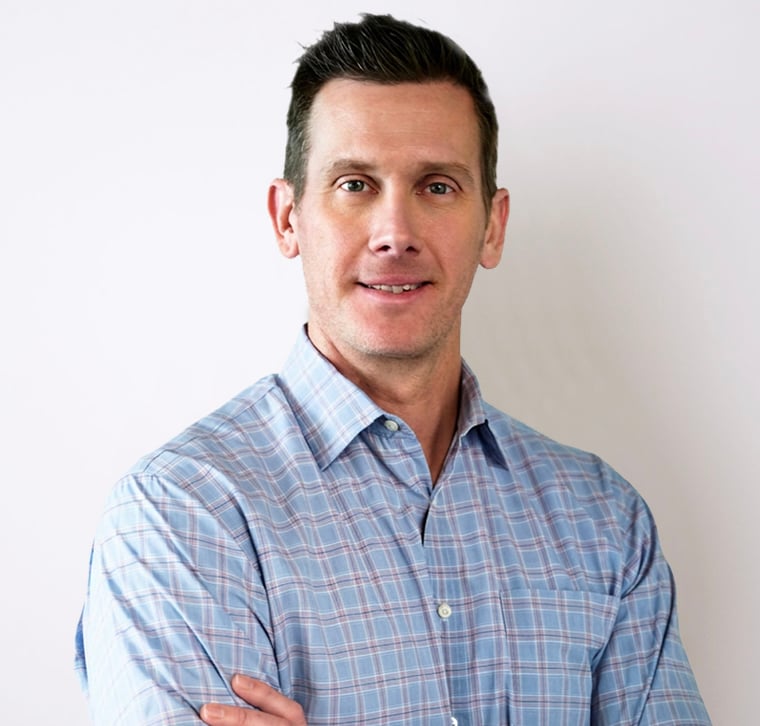 Man with short brown hair wearing a blue plaid shirt stands against a plain white background, facing the camera with a slight smile and arms crossed.