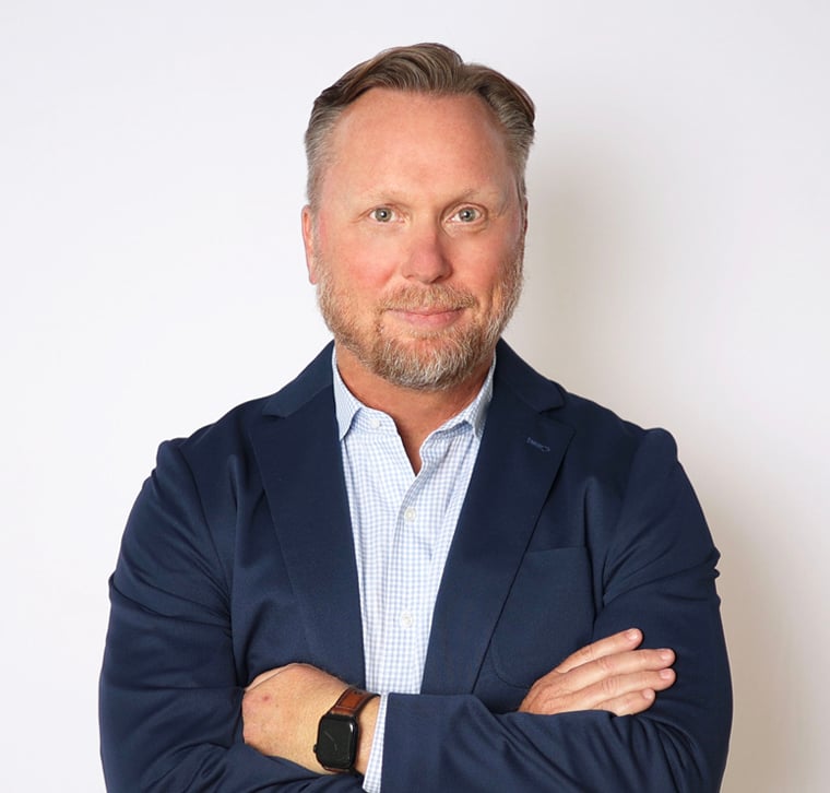 A man with light hair and a beard stands against a white background, wearing a blue blazer, a light checkered shirt, and a smartwatch, with arms crossed and a neutral expression.