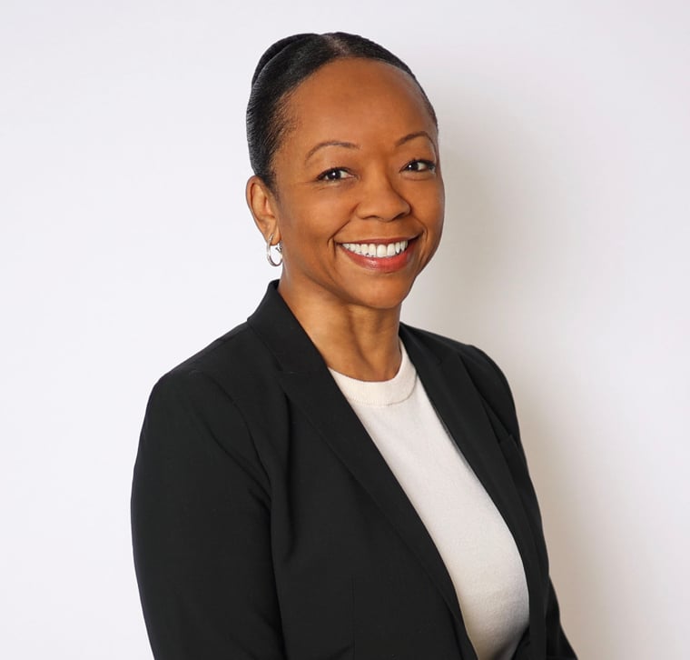 A woman in a black blazer and white top stands against a plain background, smiling with her hair pulled back.