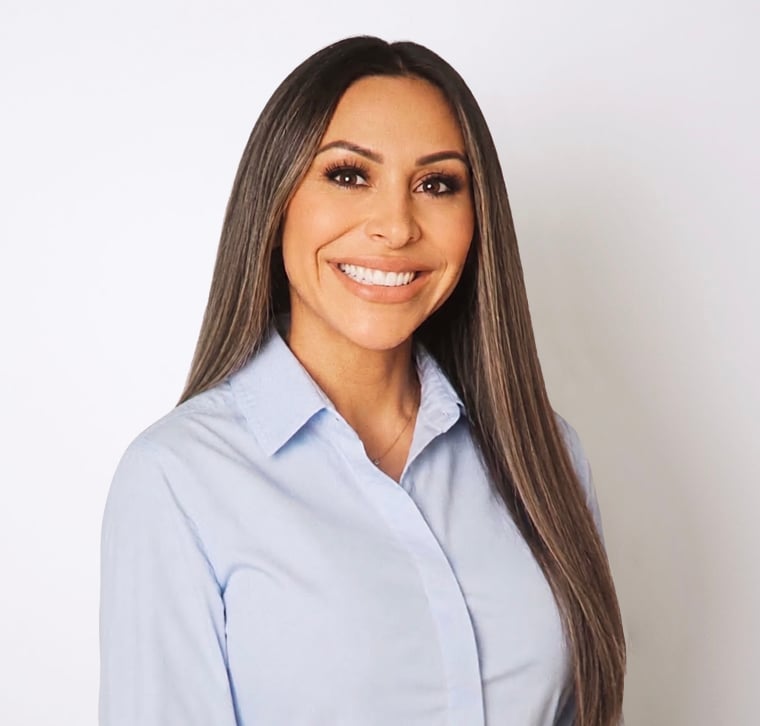 Woman with long straight brown hair wearing a light blue collared shirt, smiling, standing in front of a plain white background.
