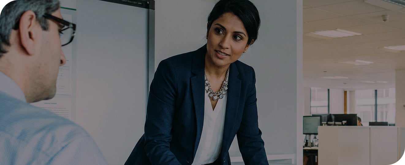 A woman in business attire stands and talks to a seated man in an office setting with desks and computers in the background.