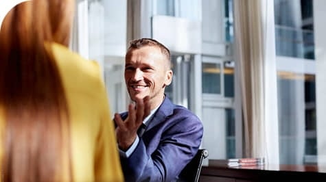 A man in a suit smiles and gestures while talking to a woman in a yellow jacket in a bright office setting.