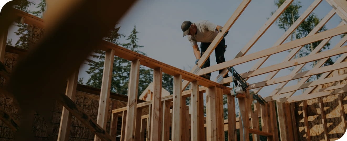 A person stands on the wooden frame of a house under construction, working outdoors with trees visible in the background.