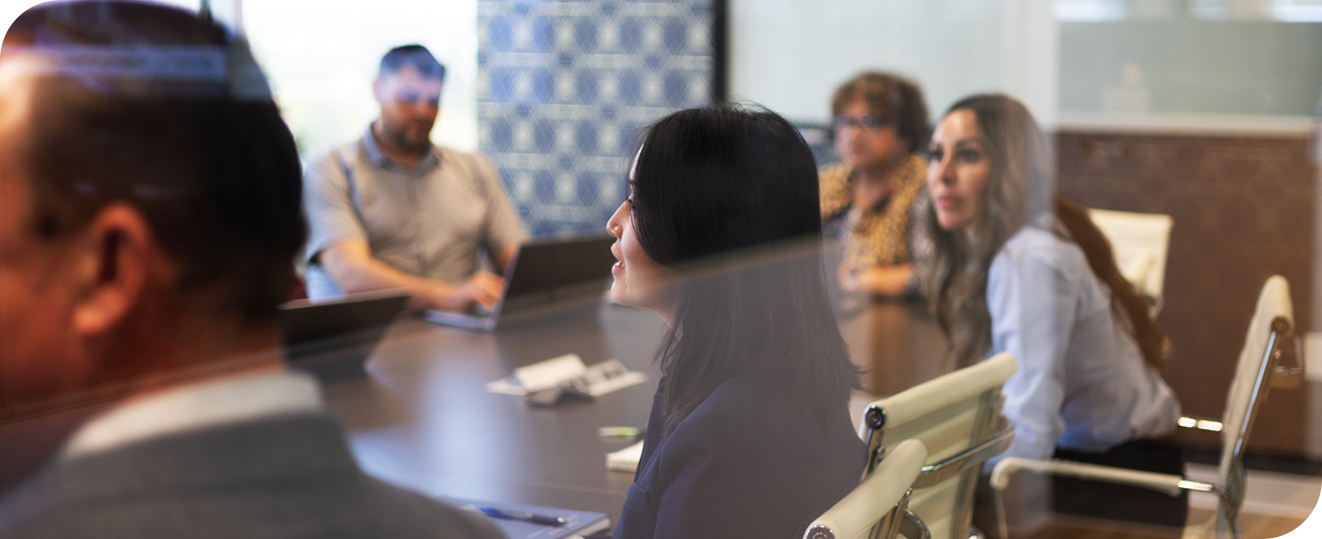 Five people sit around a conference table, some with laptops, attentively listening during a meeting in a modern office setting.
