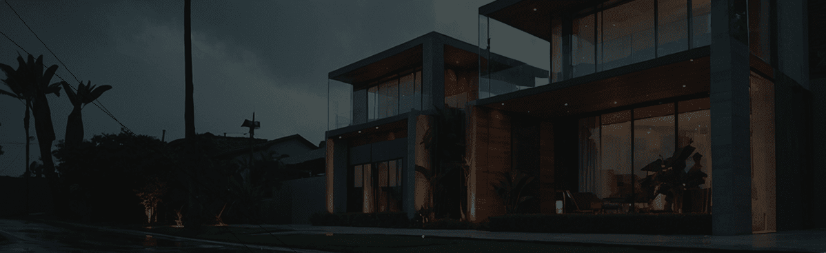 Modern two-story house with large glass windows illuminated at night, surrounded by trees and a wet driveway.