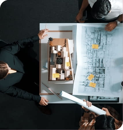 Three people review architectural blueprints and a building model on a table, with notes visible on the plans.