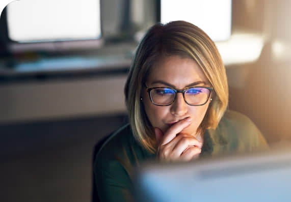 A woman wearing glasses sits at a desk, looking intently at a computer screen with her hand resting on her chin.