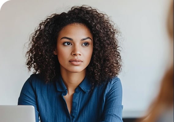 A woman with curly hair wearing a blue blouse sits at a desk, looking slightly to the side with a neutral expression.