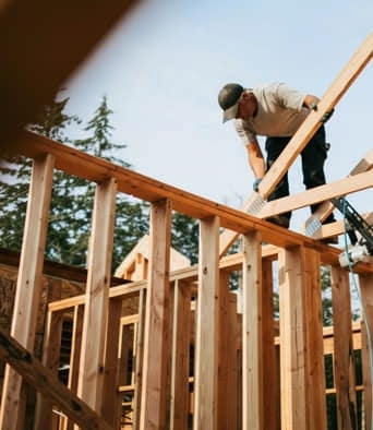 A construction worker stands on wooden framing, securing beams on a building under construction, with trees and sky in the background.