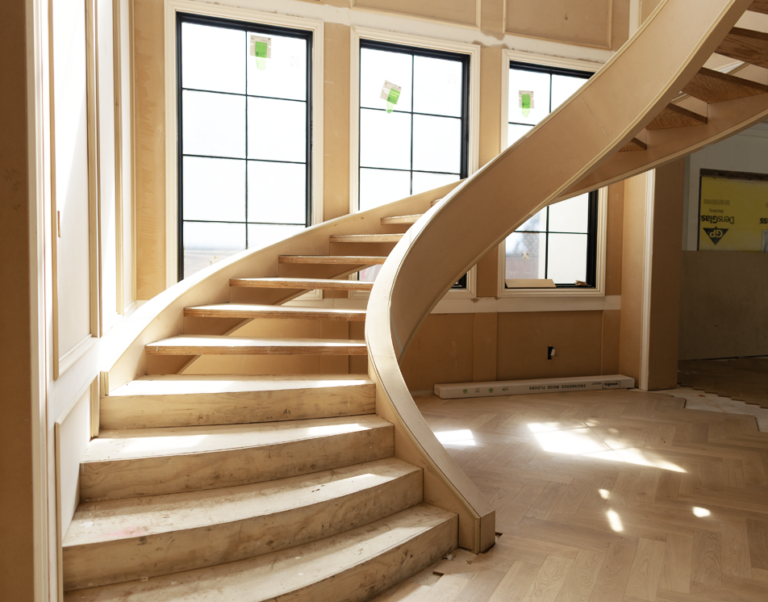 Sunlight streams through large windows onto an unfinished wooden staircase inside a building under construction or renovation.