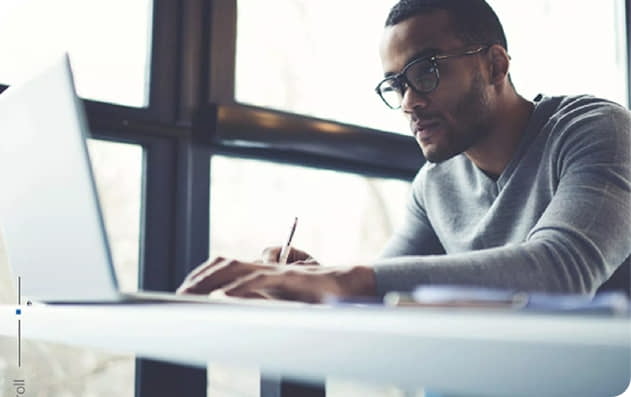 A person wearing glasses sits at a desk writing in a notebook while using a laptop, with large windows in the background.