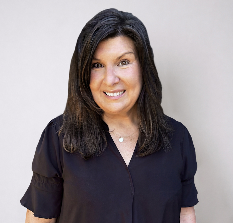 A woman with long brown hair wearing a black blouse stands smiling in front of a plain, light-colored background.