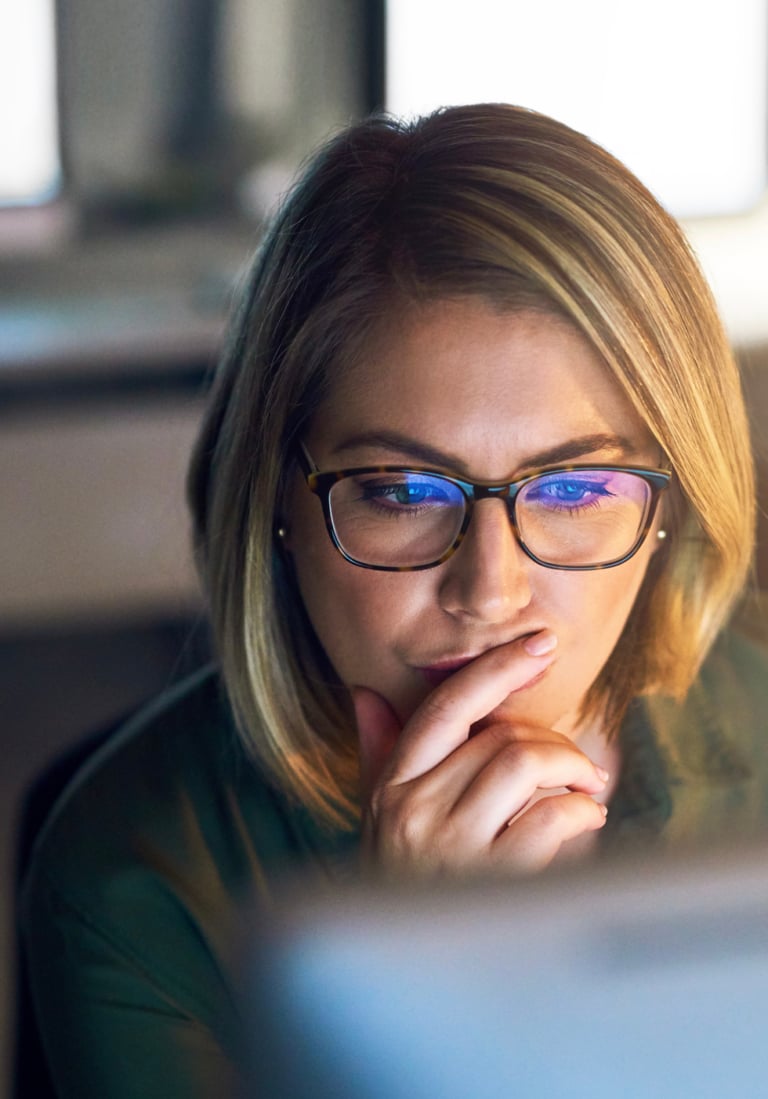 A person with glasses and shoulder-length hair looks thoughtfully at a computer screen, resting their hand on their chin.