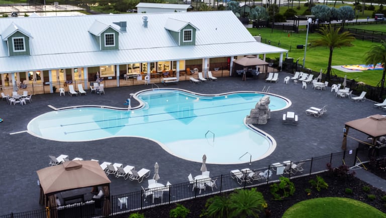 Outdoor swimming pool with lounge chairs, umbrellas, and a poolside building. The pool area is surrounded by a black fence and green lawns.