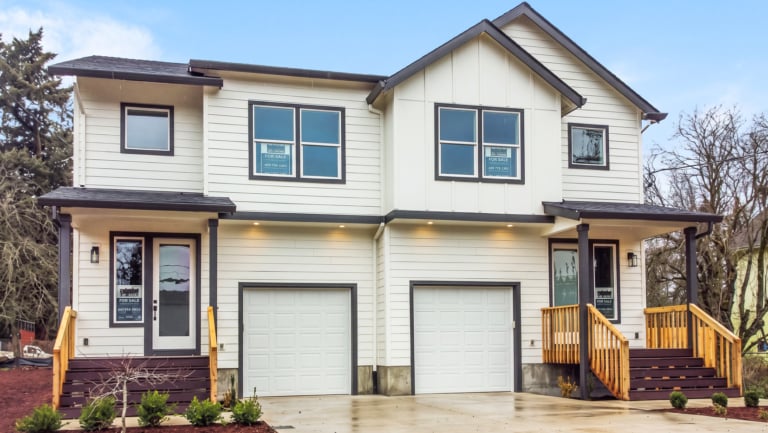 A modern two-story duplex with white siding, two garages, and wooden front porches, photographed on a clear day.