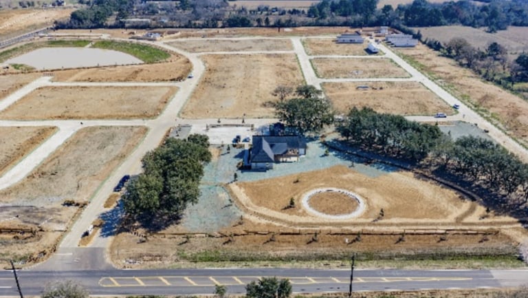 Aerial view of a residential construction site with one completed house, empty lots, partially paved roads, and surrounding trees.