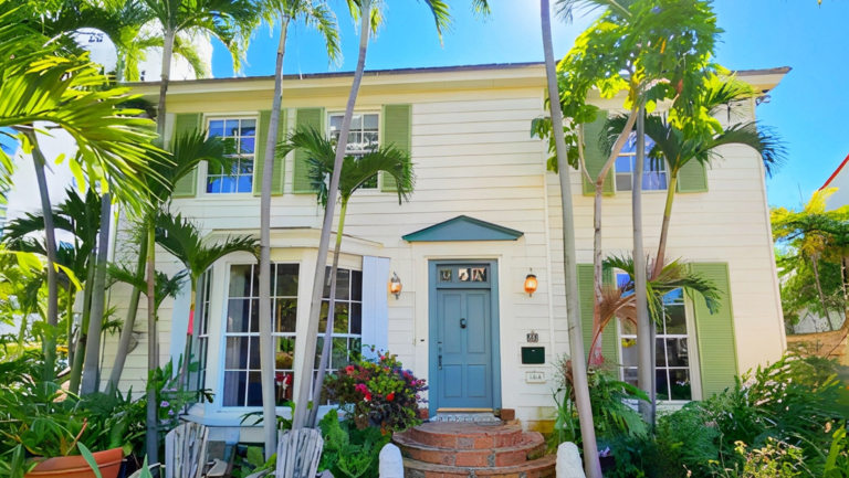 Two-story cream-colored house with green shutters, a blue front door, and surrounded by palm trees and lush plants under a clear blue sky.