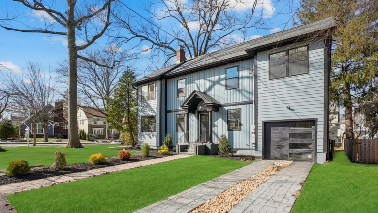Modern two-story gray house with black trim, attached garage, and landscaped yard featuring green grass and a stone walkway under a bright blue sky.