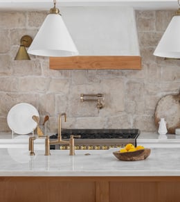 Modern kitchen with a marble countertop, brass fixtures, a stove with a pot filler, pendant lights, and a bowl of lemons, set against a stone backsplash.