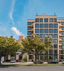 A mid-rise modern apartment building with large windows and balconies, flanked by trees and cars parked along the street under a blue sky.