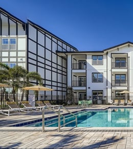 Outdoor swimming pool with metal handrails, lounge chairs, and umbrellas in front of a modern, three-story apartment building with large windows and palm trees.