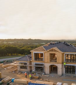 A two-story house under construction with workers on site, surrounded by building materials and green landscape in the background.