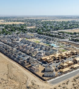 Aerial view of a suburban housing development under construction, bordered by undeveloped land, with completed homes, partially built structures, streets, and a pool area visible.