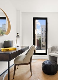 A modern home office with a desk, chair, lamp, books, round mirror, window with city view, armchair, black pouf, and cowhide rug.