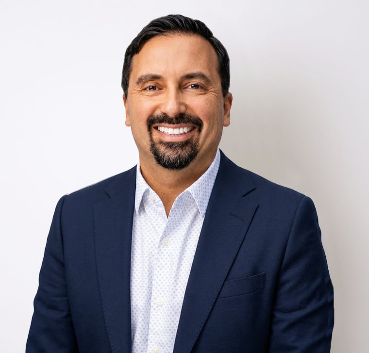 Man with dark hair, goatee, and mustache, wearing a navy blue suit jacket and a white shirt, smiling against a plain white background.